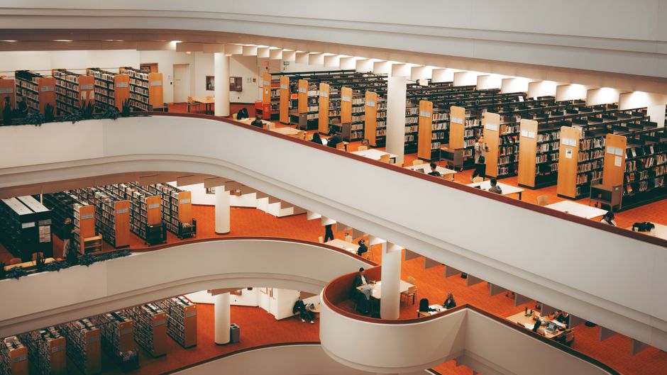 Two adults engaged in reading activities in a public library with abundant bookshelves.