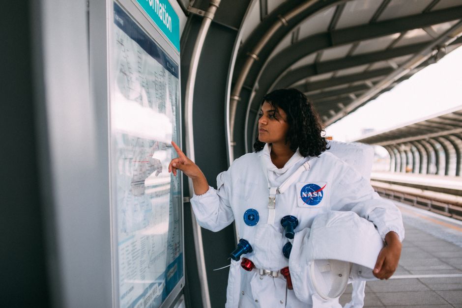 Woman dressed in NASA suit interacting with metro information display at outdoor station.